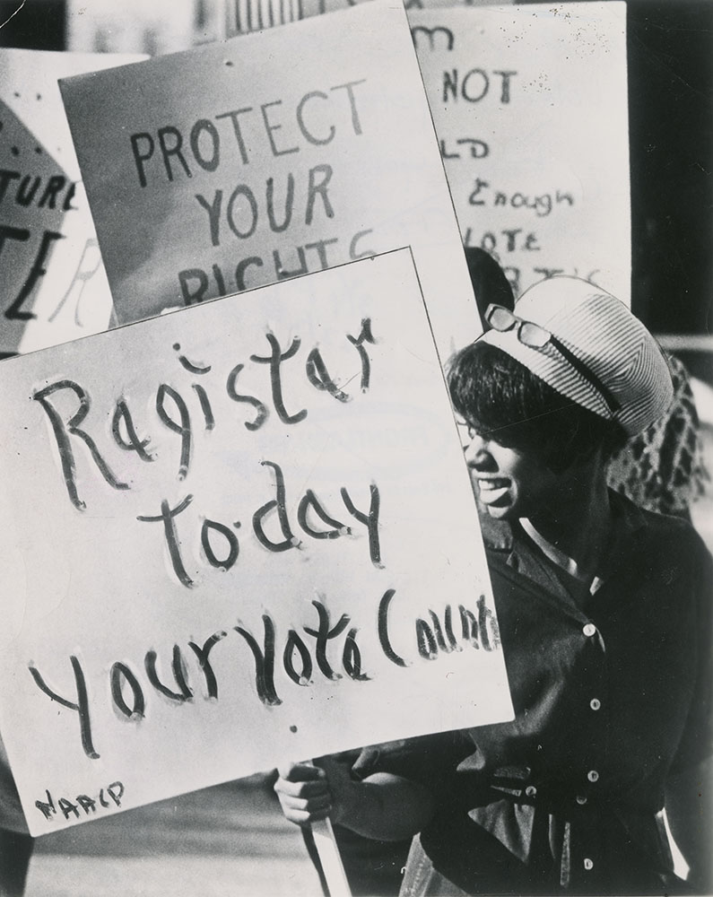 Photograph of a woman holding a sign that says 'register today your vote counts'