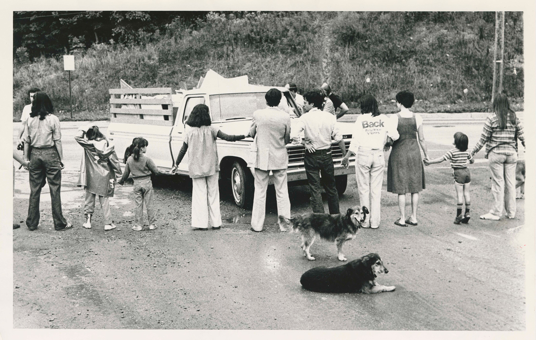 People of all ages (kids and adults) forming a human chain, blocking a pickup truck carrying landfill.