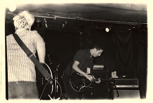 Alexander Dunham, in a black short sleeved shirt plays a dark colored hollowbody guitar as he bends over the instrument.