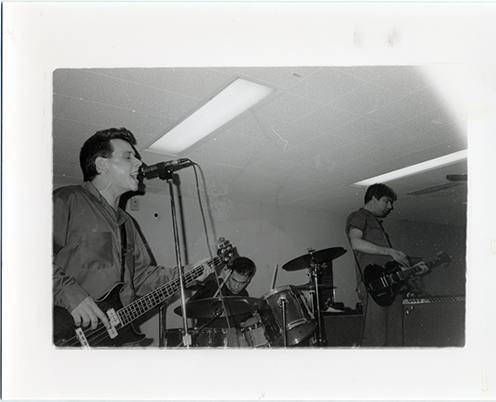 Black and white photograph of three young men playing guitar, bass and drums in a small room.