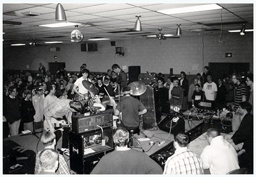 A black and white photograph of a band composed of four young men playing guitars and drums on a small elevated stage, surrounded on all sides by audience members.