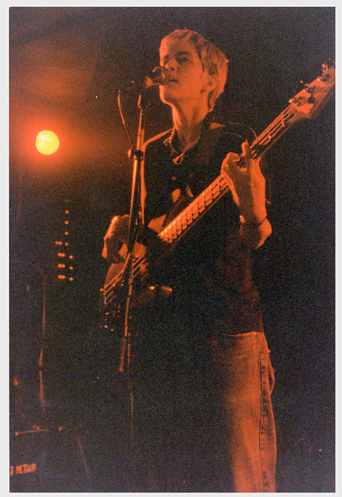 A photograph of Johanna Claasen, with short light-colored hair, black button-down shirt and baggy jeans plays a black bass guitar and sings onstage.