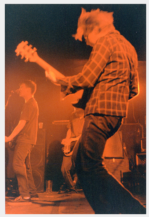 A photograph of a man in a flannel shirt plays guitar in the foreground while a man plays guitars and sings with a scrunched face onstage.