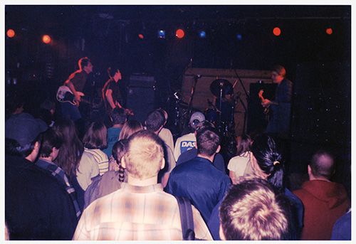 A color photograph of a four-piece band onstage looking toward their drummer in front of four rows of audience members standing in front of the stage.