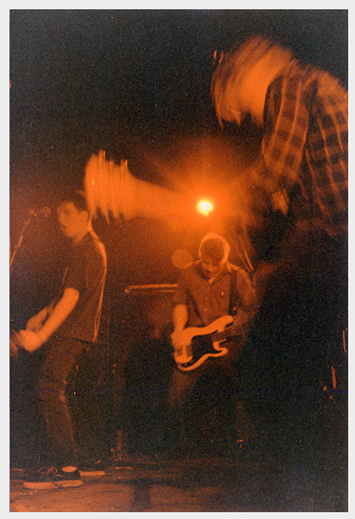 A photograph of three young men playing guitars onstage, blurred by their motion.