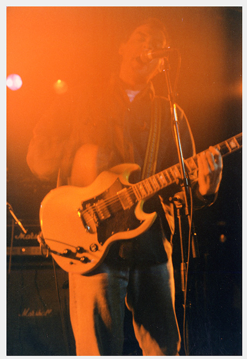 A photograph of Marc Nelson, with short brown hair wearing an overshirt and jeans playing an SG-shaped guitar plays and sings onstage obsacured by stage lights.