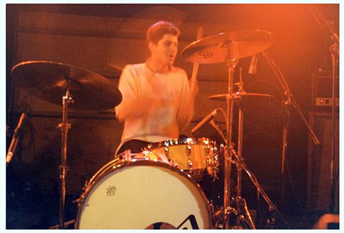 Wearing a plain white t-shirt, Ryan Nelson plays drums onstage in a motion-blurred photograph.