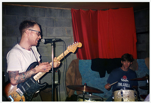 A poster with a black and white fisheye-lens photo of a drumset and other instruments in a recording studio, the photo is processed to be mostly blue and black and there is bold elongated yellow text.