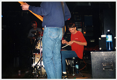 A poster with a black and white fisheye-lens photo of a drumset and other instruments in a recording studio, the photo is processed to be mostly blue and black and there is bold elongated yellow text.