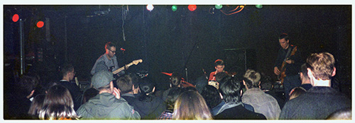 A poster with a black and white fisheye-lens photo of a drumset and other instruments in a recording studio, the photo is processed to be mostly blue and black and there is bold elongated yellow text.